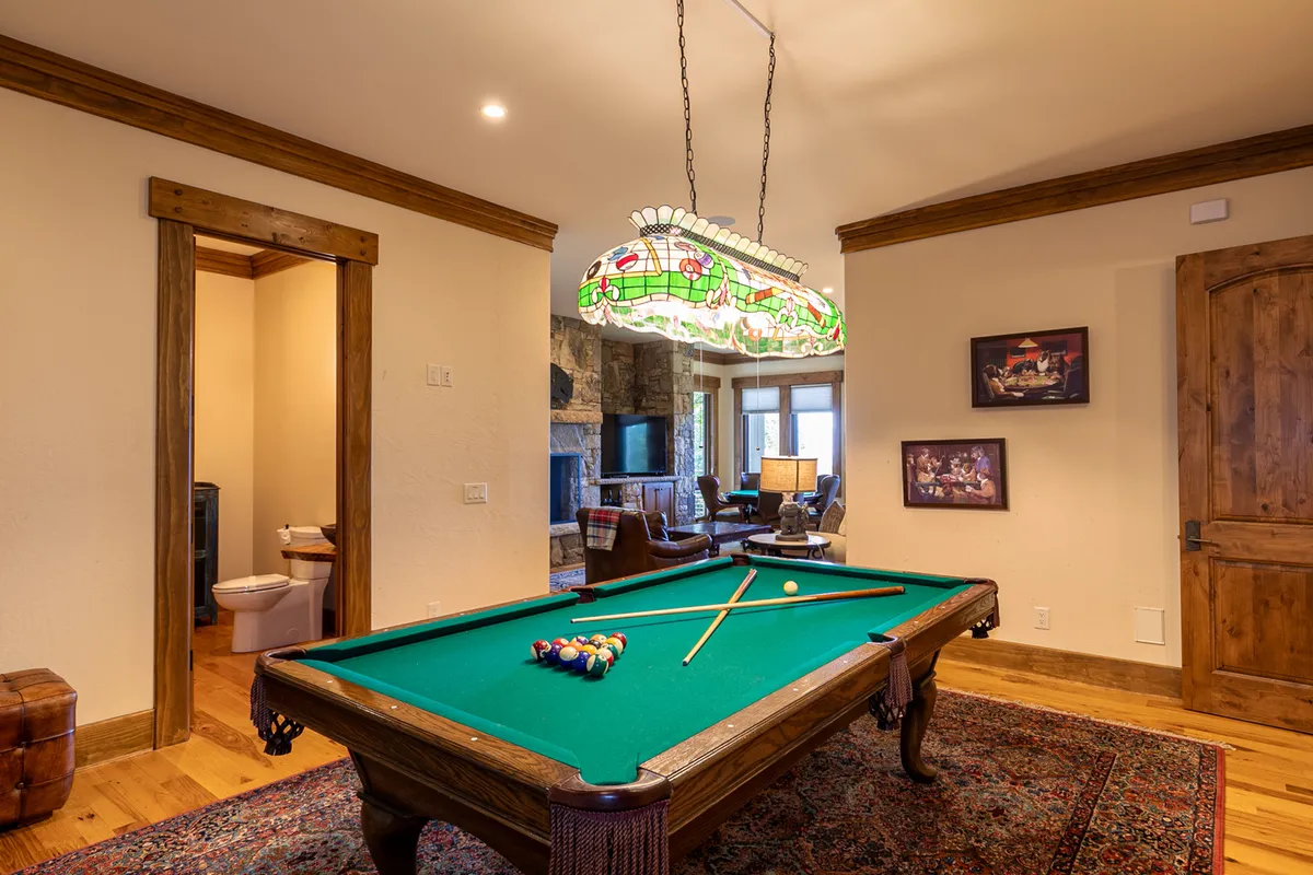 This image features a stylish game room anchored by a classic pool table under a stained glass light fixture. The space also includes a cozy fireplace and a comfortable seating area, creating an inviting environment for both spirited competition and relaxation.