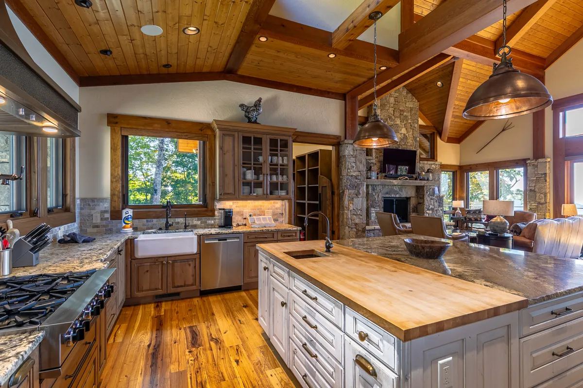 A wide view of the kitchen and adjacent area, featuring a large kitchen island, stone fireplace, wooden beams, and generous windows. The setup encourages both cooking and social interaction in a warm, cozy environment.