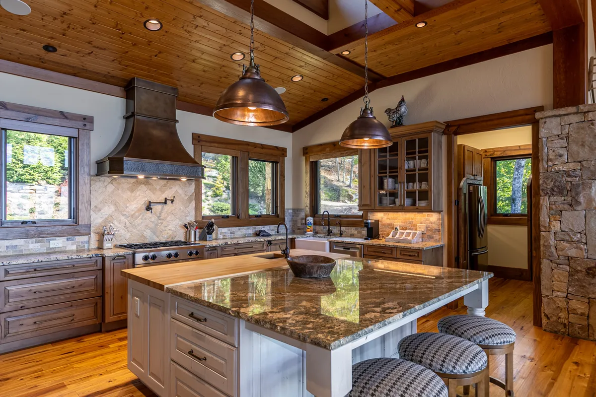 A gourmet kitchen featuring a large kitchen island with bar stools, copper pendant lights, stainless steel appliances, and a natural stone backsplash. The space is warmly lit and designed to accommodate multiple people for both cooking and socializing.