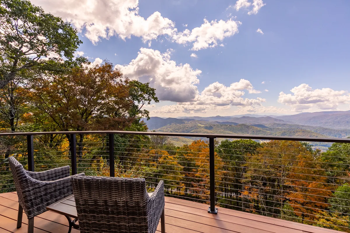 Two comfortable chairs are set on a viewing spot overlooking a breathtaking mountain vista under a sky painted with soft clouds. The mood is quiet and contemplative, inviting guests to relax, read, or share a glass of wine while soaking in the panoramic view.