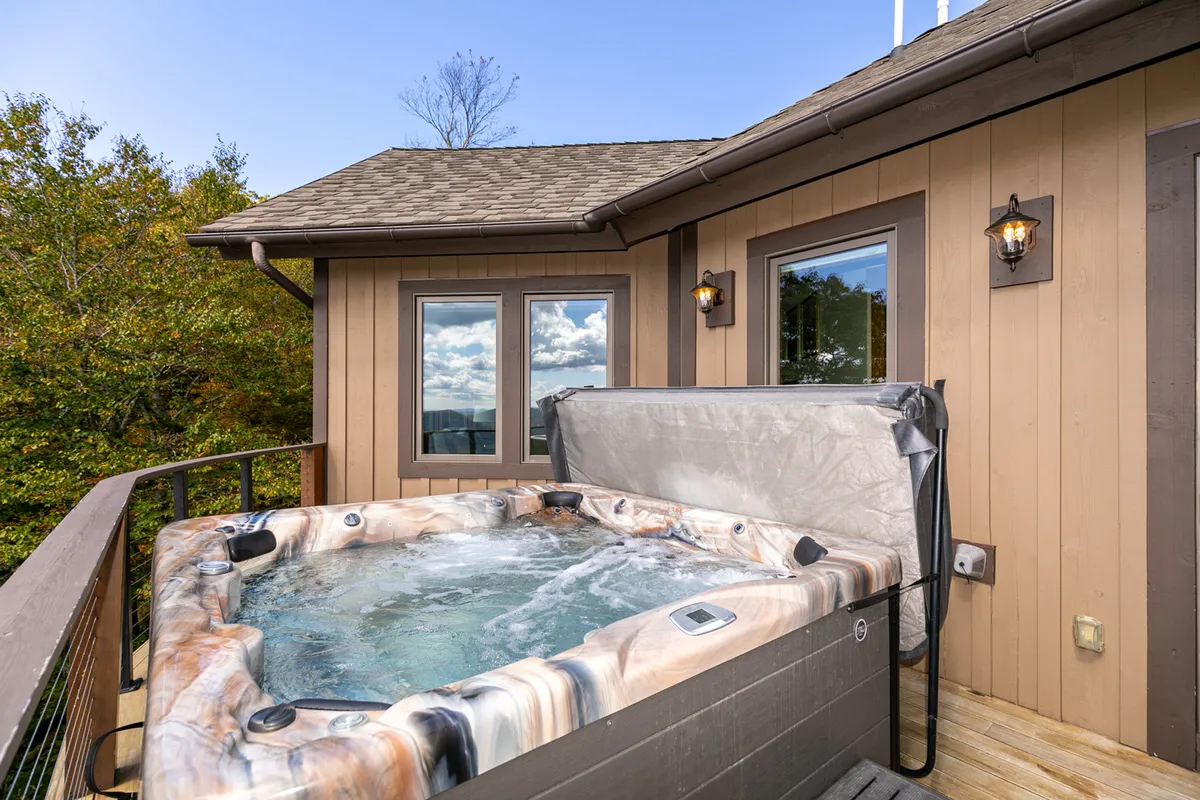 A cozy outdoor hot tub nestled among trees, with the property’s rustic exterior and large windows visible nearby. The scene conveys privacy and relaxation, suggesting the perfect spot to unwind after a day of mountain exploration, surrounded by nature’s tranquility.