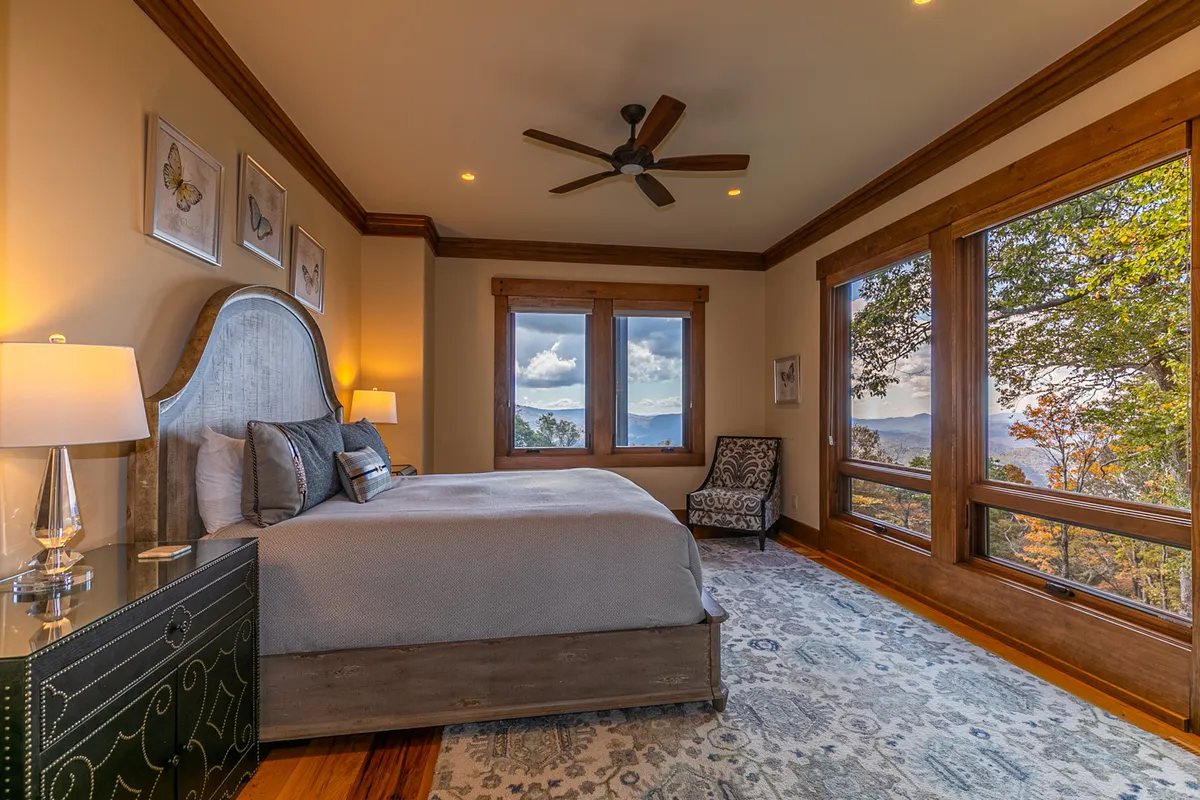 A serene, sunlit bedroom with a large bed draped in plush bedding, decorative chair by the window, and a nightstand with a lamp. Expansive windows reveal tranquil mountain views, creating an atmosphere of peace and relaxation.