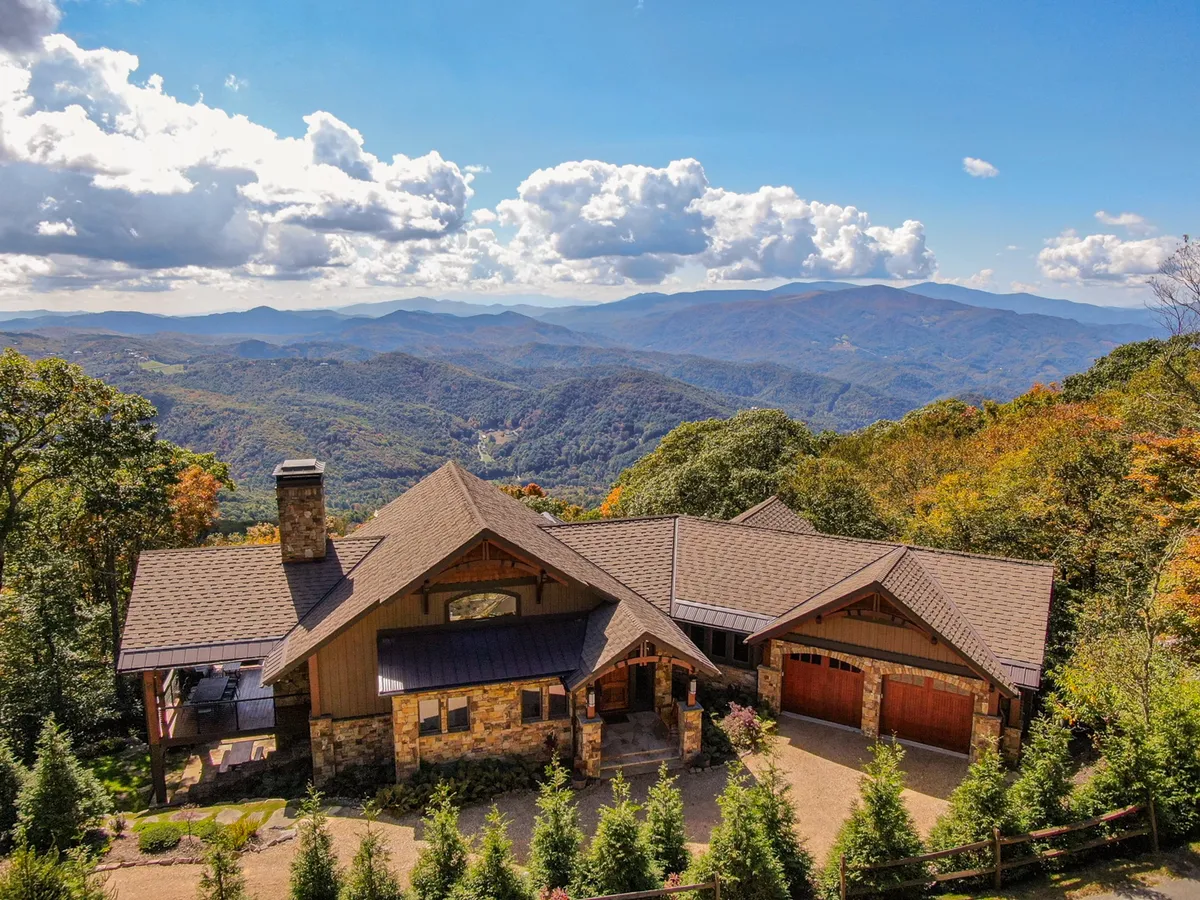 The image showcases a stunning mountain home with a stone facade, perched amidst lush trees and overlooking sweeping 180-degree mountain vistas beneath a clear blue sky. The expansive deck hints at outdoor relaxation, while the natural beauty of the surrounding landscape offers a tranquil, inviting atmosphere.