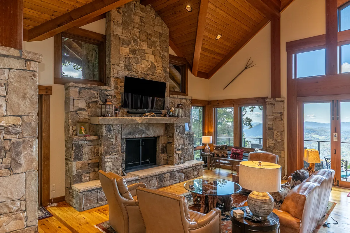 A warm, welcoming living area featuring a grand stone fireplace, expansive windows framing wide mountain vistas, plush seating, and rustic exposed wooden beams overhead. Natural light pours in, highlighting the open, airy ambiance ideal for gatherings.