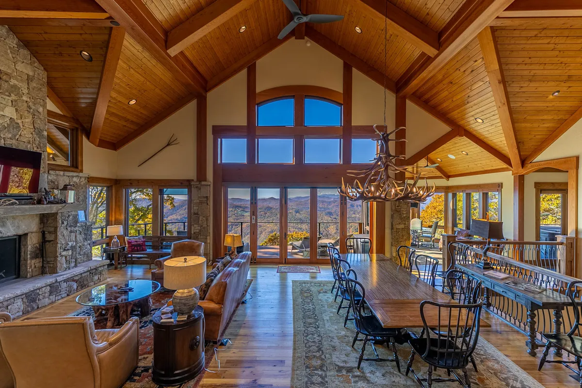 A spacious, open-concept living and dining area with vaulted wooden ceilings, a striking stone fireplace, and a sleek long dining table beneath a stylish chandelier. Massive windows bathe the room in light and offer unobstructed sunset views of the mountains.