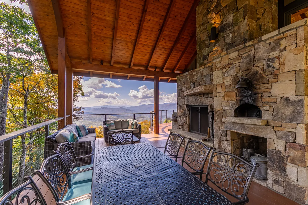 This exterior image showcases a spacious outdoor dining area with a large table, comfortable seating, a stone fireplace, and panoramic mountain views. The scene is set for sunset, emanating a welcoming and convivial atmosphere perfect for group celebrations or al fresco meals.
