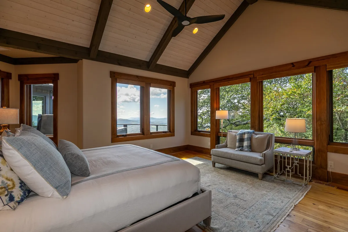 A beautifully appointed bedroom featuring a large bed with decorative pillows, a comfortable armchair positioned by expansive windows, and a stylish side table. The room is bathed in natural light with views of the mountains, offering a sanctuary-like feel.