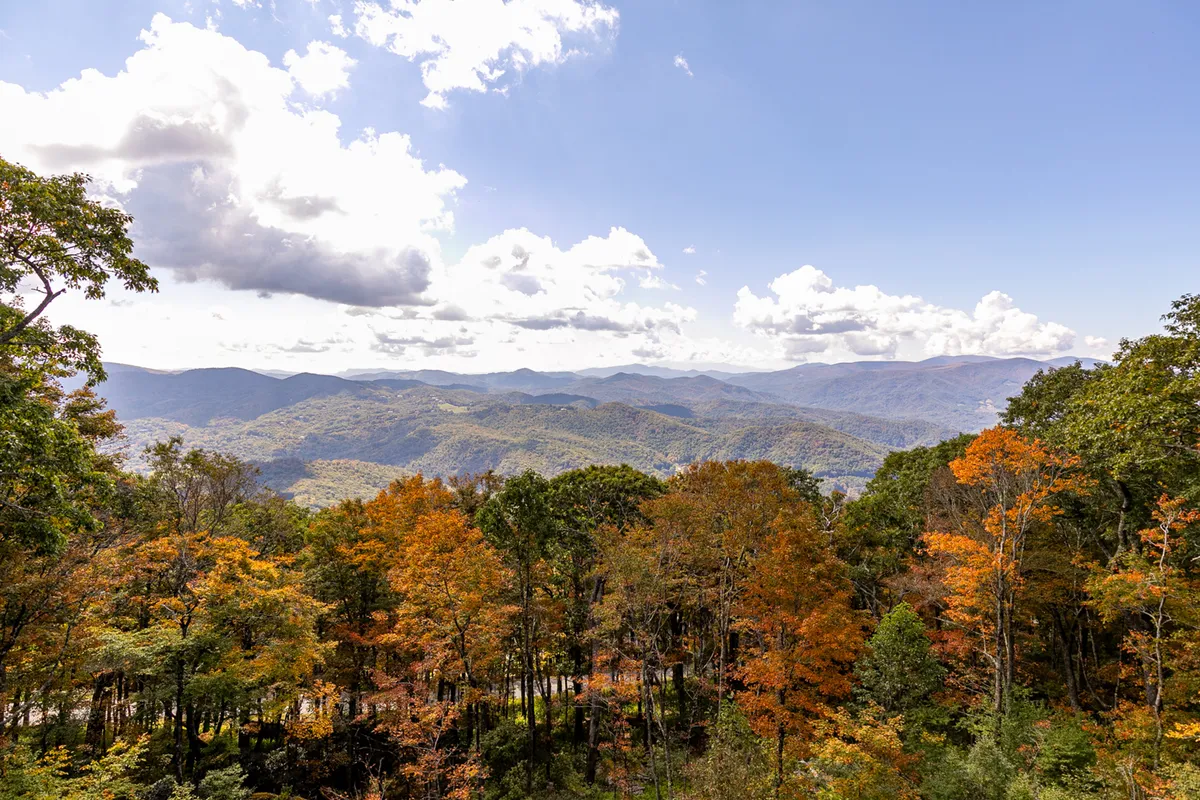 A panoramic view of a majestic mountain range bathed in autumn hues, with rolling clouds and vibrant foliage creating a dramatic and inviting outdoor scene.